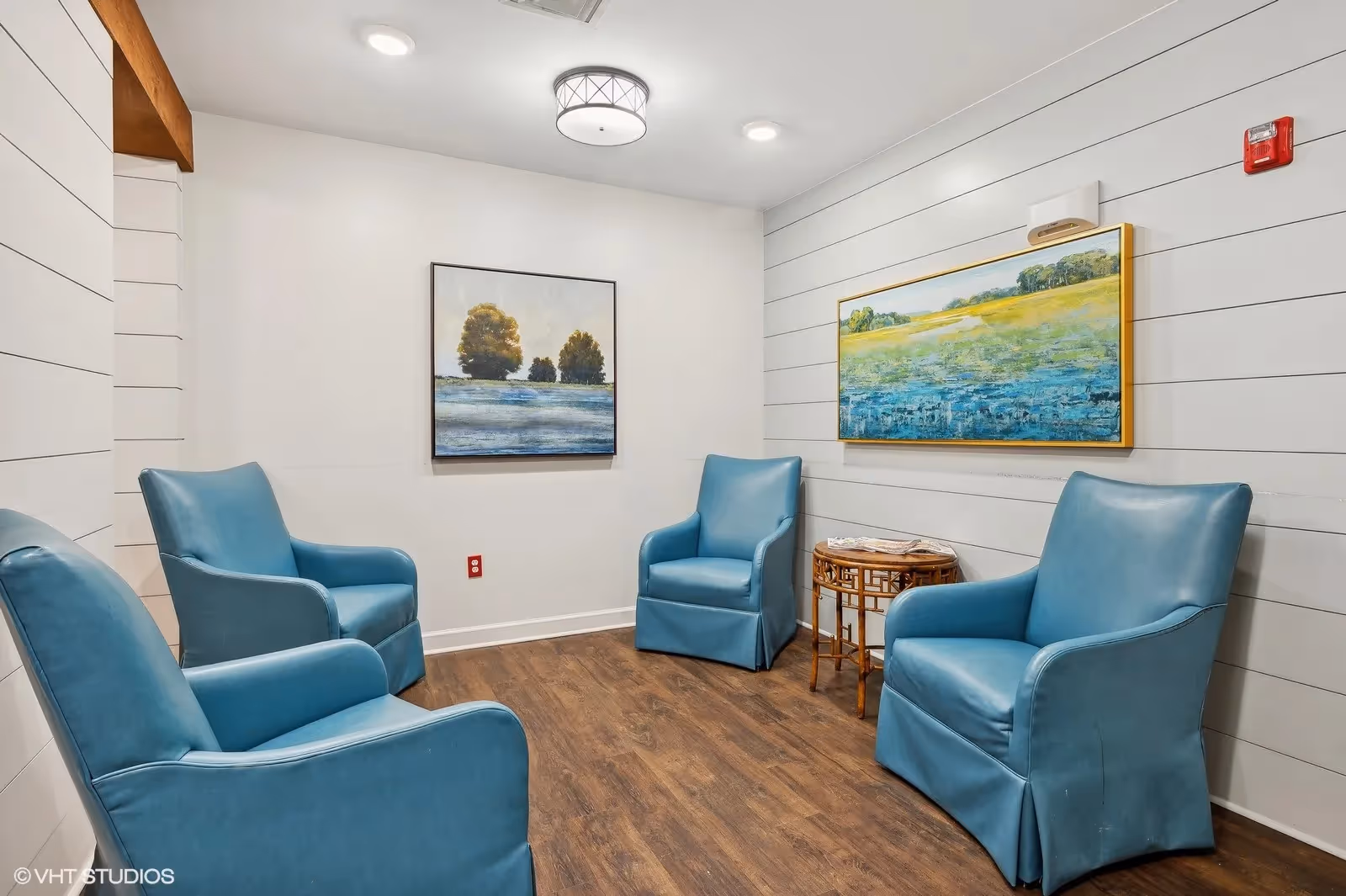 Small seating lounge with four blue upholstered chairs around a wicker side table and landscape paintings on white shiplap walls.