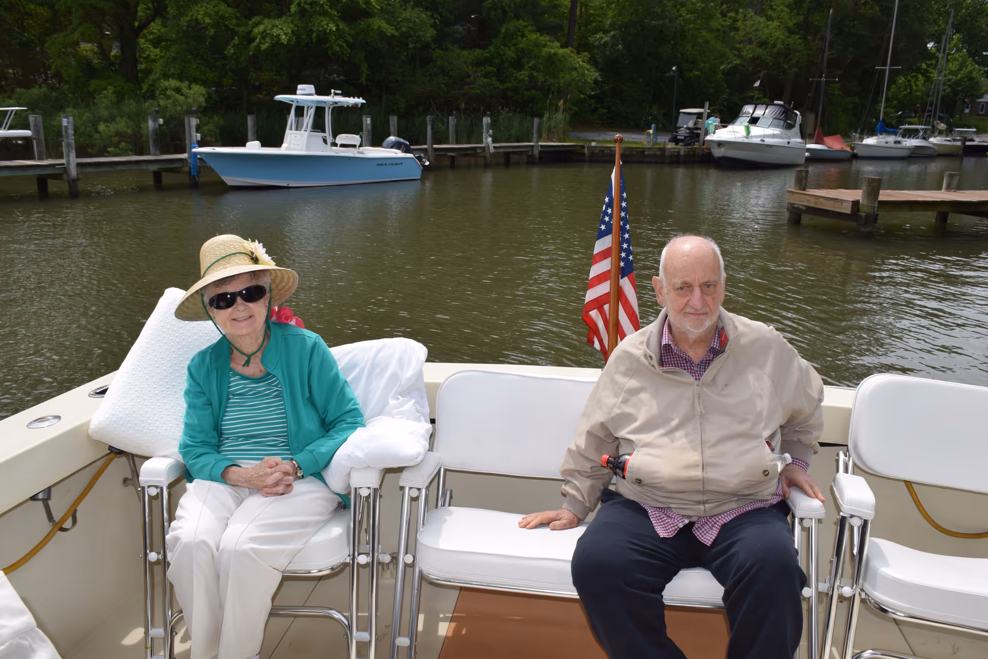 An elderly woman wearing a green jacket, striped shirt, white pants, sunglasses, and a sun hat sits on a white chair on a boat. Next to her, an elderly man in a beige jacket and dark pants sits on a white bench. Behind them is a body of water with several boats docked and an American flag mounted on the boat.