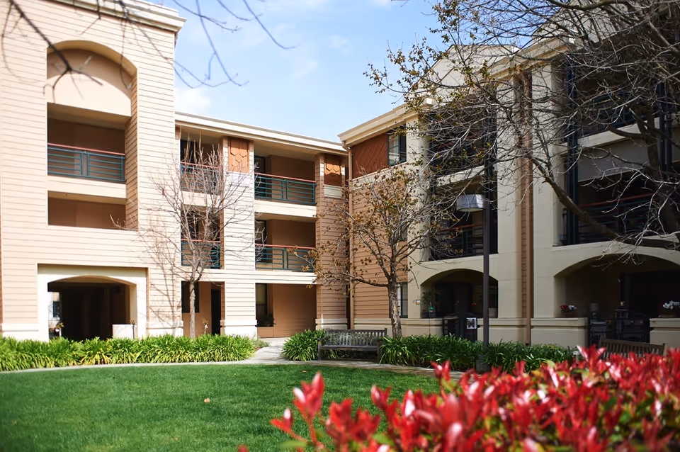 Exterior view of a multi-story senior living facility building with beige and brown walls, balconies with green railings, leafless trees, green grass, and red flowering plants in the foreground under a clear sky.