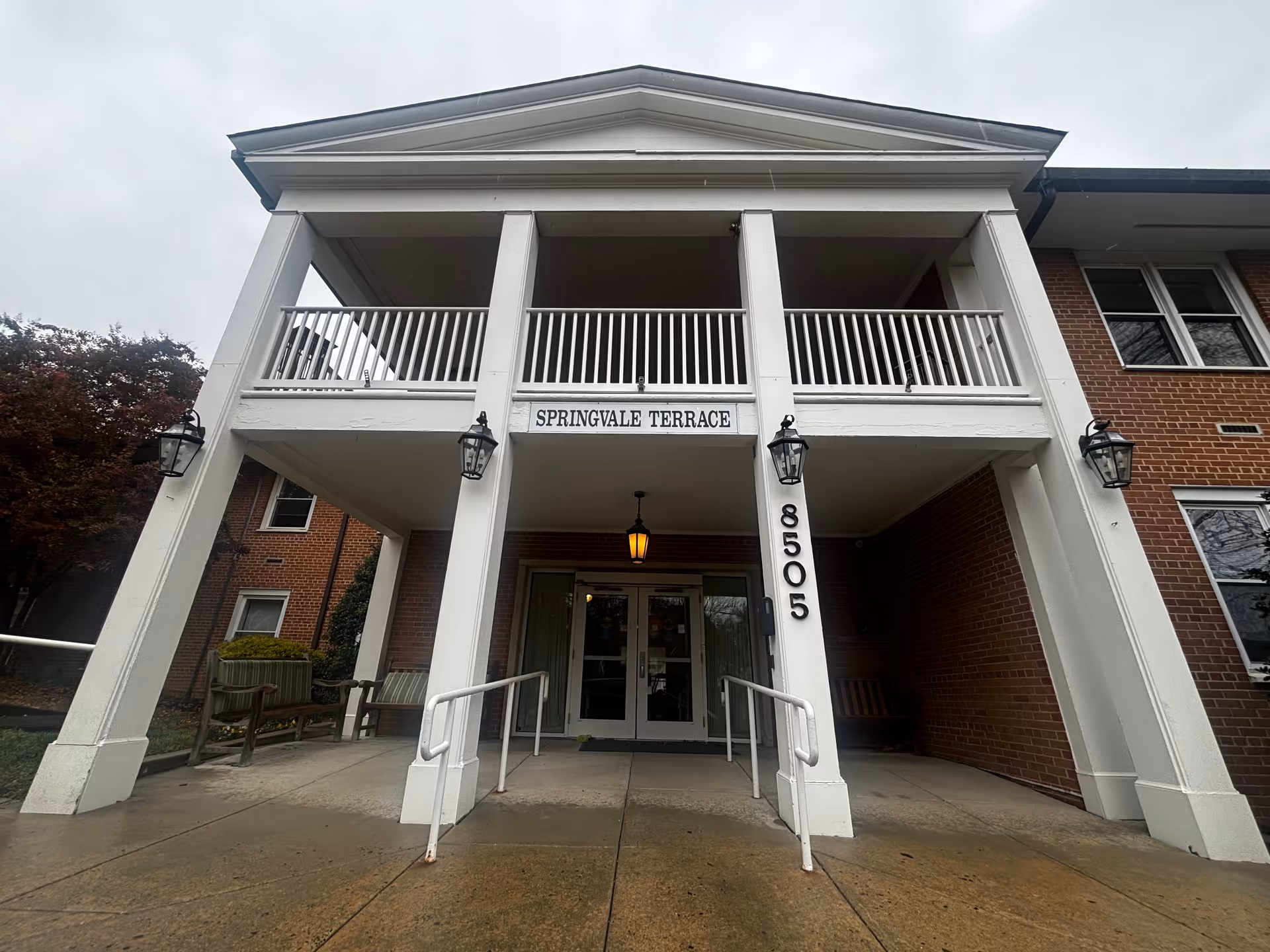 Front entrance of a brick building with white columns and railings. The sign above the entrance reads 'SPRINGVALE TERRACE' and the building number '8505' is displayed vertically on one of the columns. There are benches on either side of the entrance and outdoor lantern-style lights mounted on the columns.