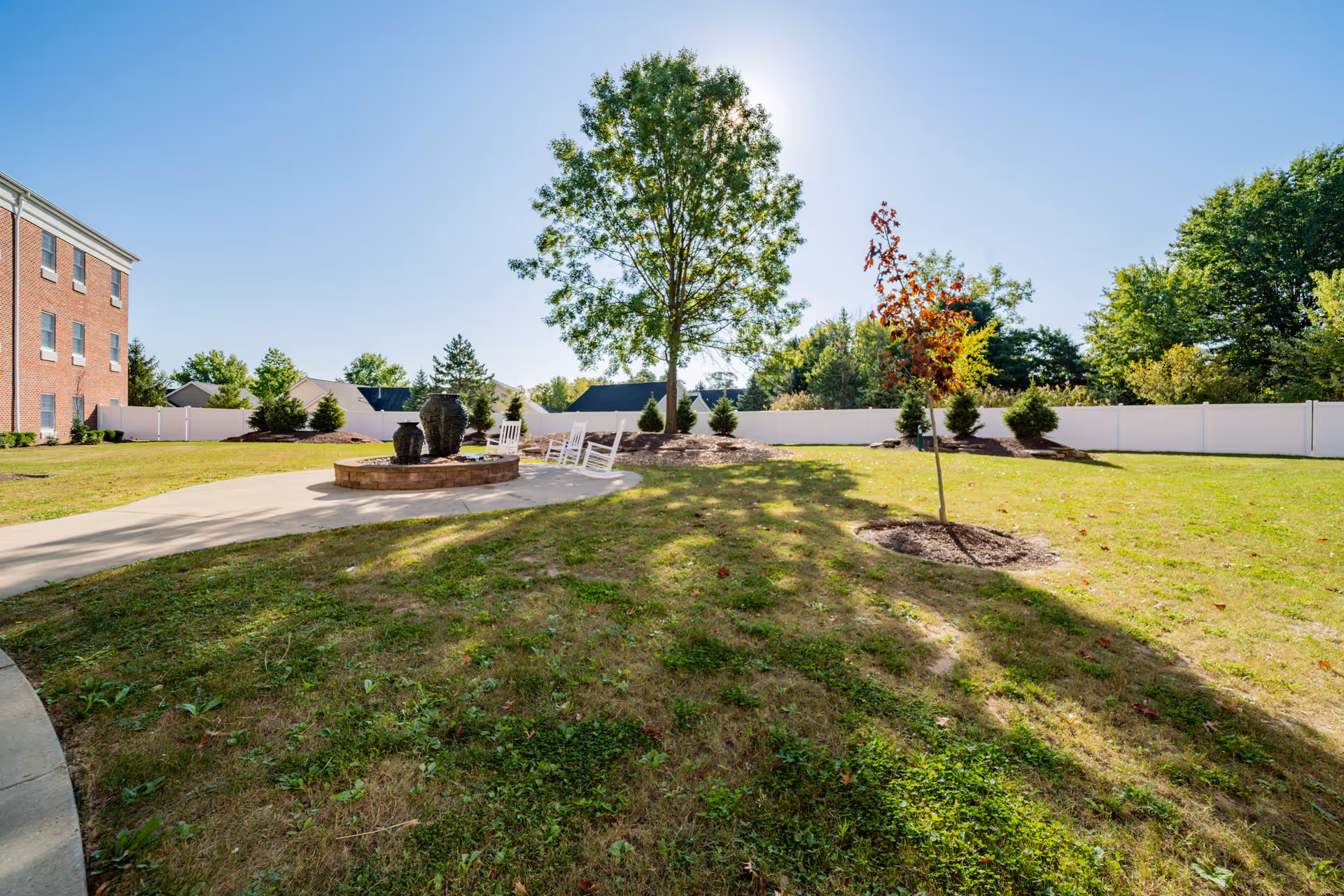 Outdoor garden area with a concrete pathway leading to a circular brick fire pit surrounded by white rocking chairs. There are several trees and shrubs planted around the grassy lawn, with a white fence enclosing the area. A red brick building is visible on the left side under a clear blue sky.