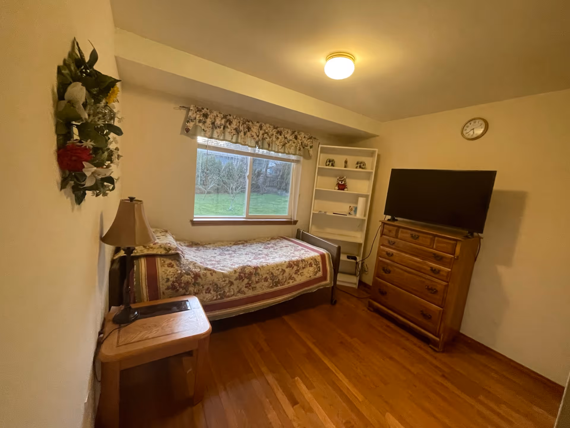 A small bedroom with a single bed covered in a floral bedspread, a wooden nightstand with a lamp, a window with floral curtains showing a green lawn outside, a white bookshelf with decorative items, a wooden dresser with a flat-screen TV on top, and a clock on the wall.