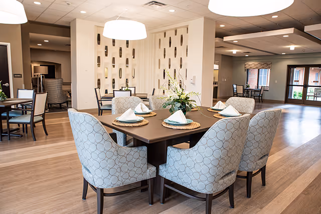 A spacious and well-lit dining area with a square table set for six people. The table has woven placemats, white folded napkins, and a floral centerpiece. The room features modern decor with patterned upholstered chairs, wooden flooring, and a decorative partition wall. Additional seating areas and large windows are visible in the background.