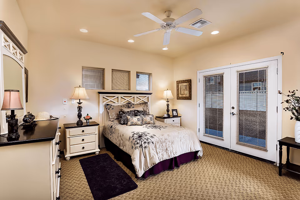 Well-lit decorated bedroom with a bed, nightstands, dresser, lamps, a ceiling fan, and French doors.