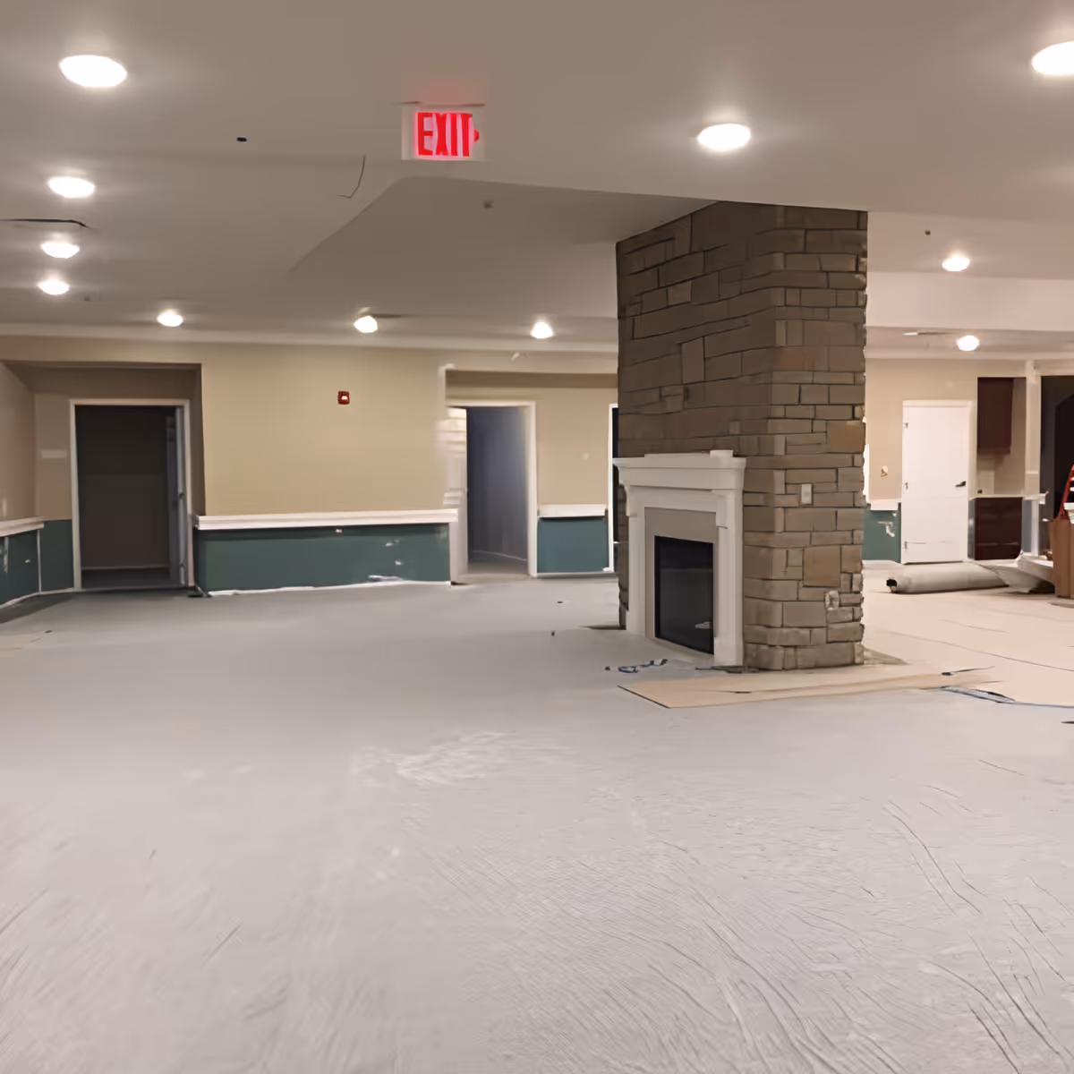 An empty interior room under renovation with beige walls, green lower wall panels, a stone fireplace in the center, and multiple doorways leading to other rooms. The floor is covered with protective material and there are ceiling lights and an illuminated exit sign.