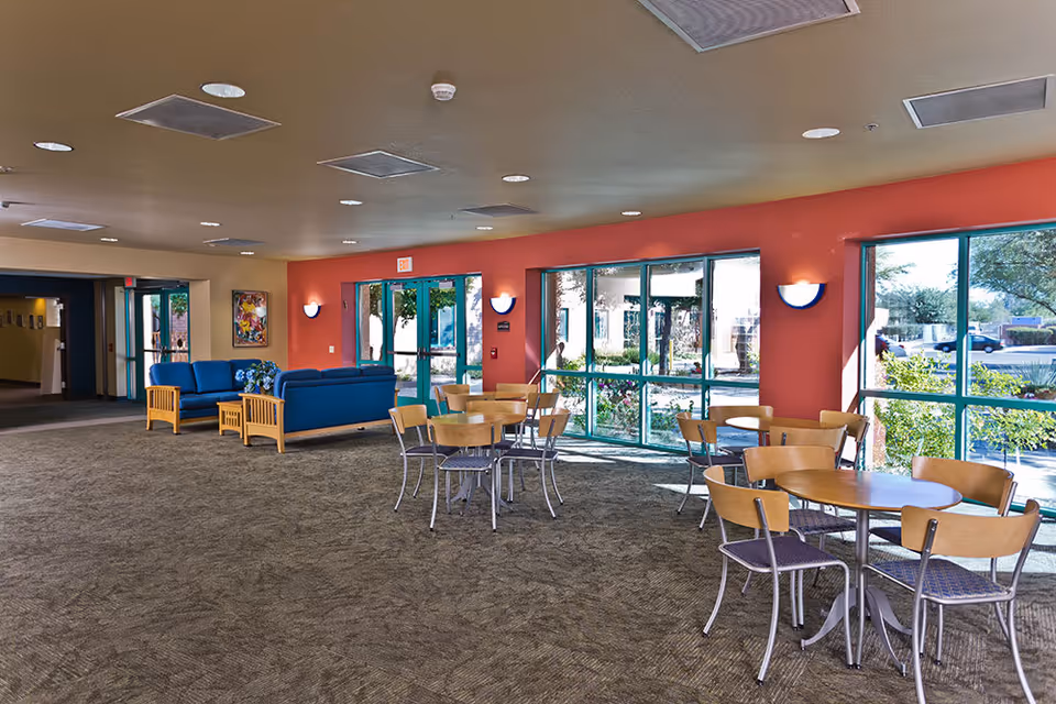 A spacious senior living community common area with several round tables and chairs arranged near large windows that let in natural light. There are two blue sofas with wooden side tables against a red accent wall. The room has a carpeted floor and ceiling lights, with doors leading outside and to other parts of the facility.