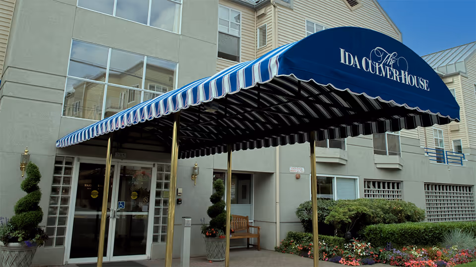 Entrance of The Ida Culver House Ravenna with a blue and white striped awning displaying the facility name. The building exterior is light-colored with large windows, potted plants, and a bench near the entrance.