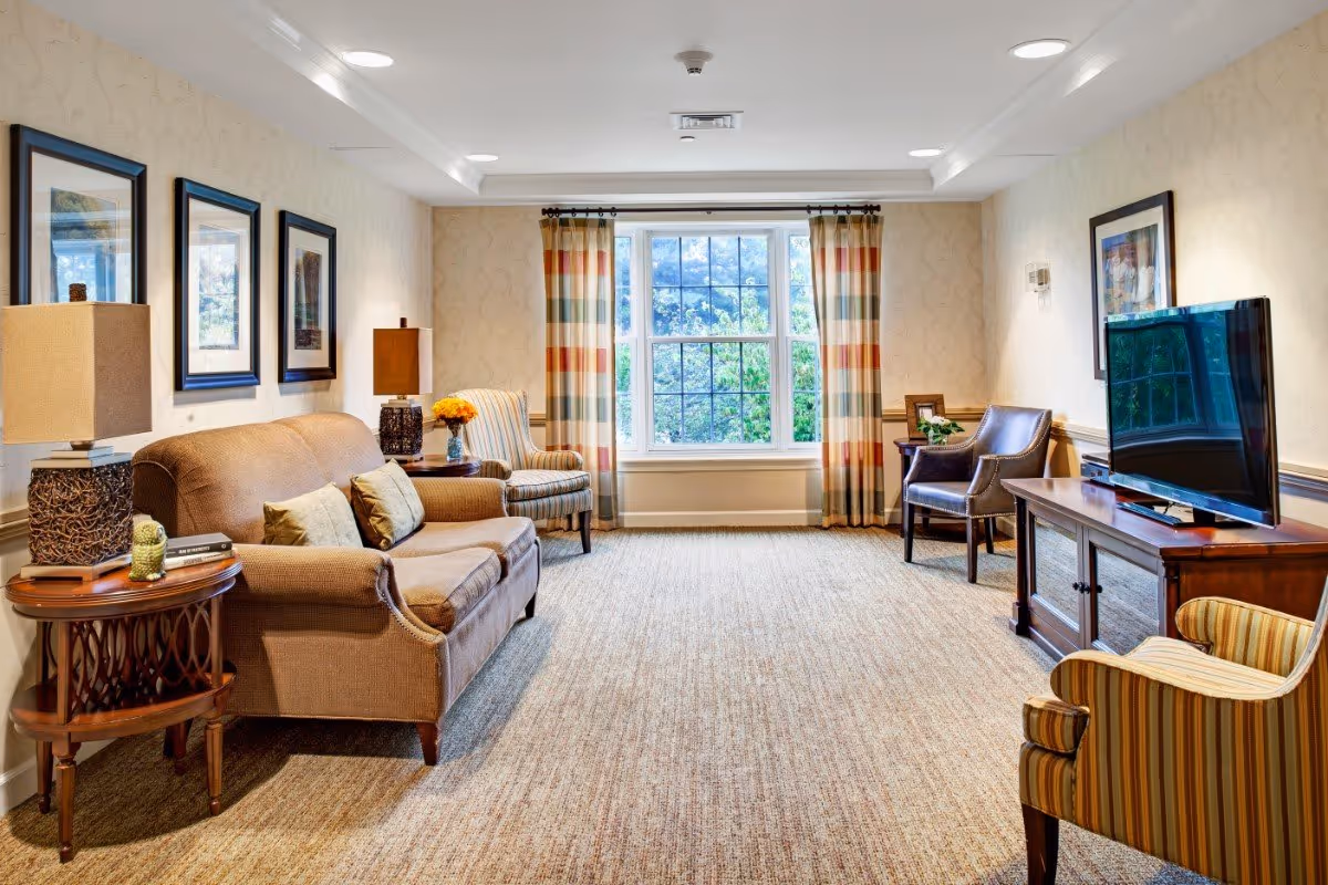 A cozy living room in a senior living facility with a brown sofa, two armchairs, a wooden TV stand with a flat-screen television, two side tables with lamps, framed artwork on the walls, and a large window with striped curtains letting in natural light.