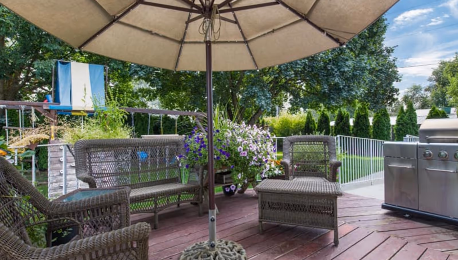 Outdoor patio area with wicker chairs and a loveseat arranged around a large beige umbrella. There is a stainless steel grill on the right side and a flower pot with purple and white flowers in the background. Trees and greenery surround the patio, and a playground structure is visible in the distance.