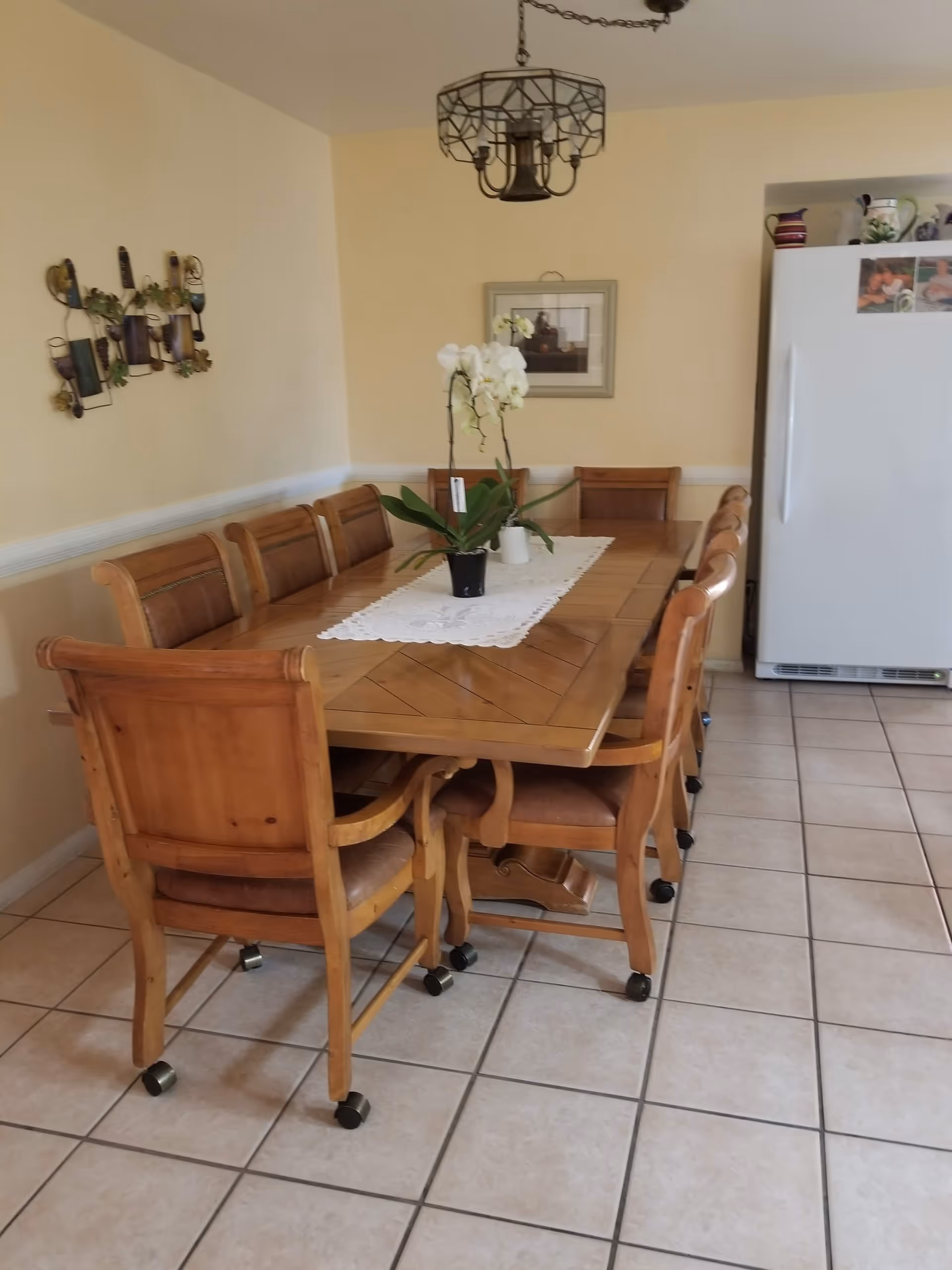 A dining room with a long wooden table surrounded by eight wooden chairs with leather seats on wheels. A white lace table runner with a potted white orchid is centered on the table. The room has tiled flooring, light yellow walls, a decorative wall hanging, a framed picture, a hanging light fixture, and a white refrigerator in the corner with photos and decorative items on top.
