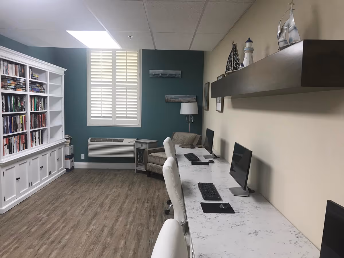 A quiet room with a long white marble-patterned desk holding three computer monitors, keyboards, and mice. There are three white office chairs in front of the desk. On the left side of the room is a large white bookshelf filled with books. The back wall is painted teal with a window featuring white plantation shutters and an air conditioning unit below it. A beige armchair and small side table with a lamp are positioned near the window. A wooden shelf above the desk holds decorative items including model sailboats and a lighthouse figurine. The floor is wood-patterned.