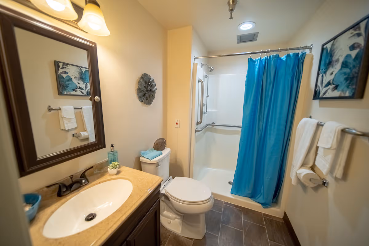 Compact bathroom with a sink and mirror, toilet, and a shower with a blue curtain and grab bars.