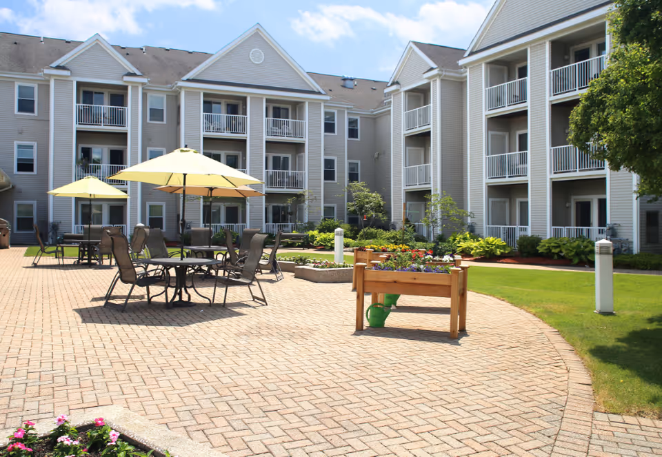 Sunny courtyard with patio tables and umbrellas, raised planters, and a multi-story residential building with balconies.
