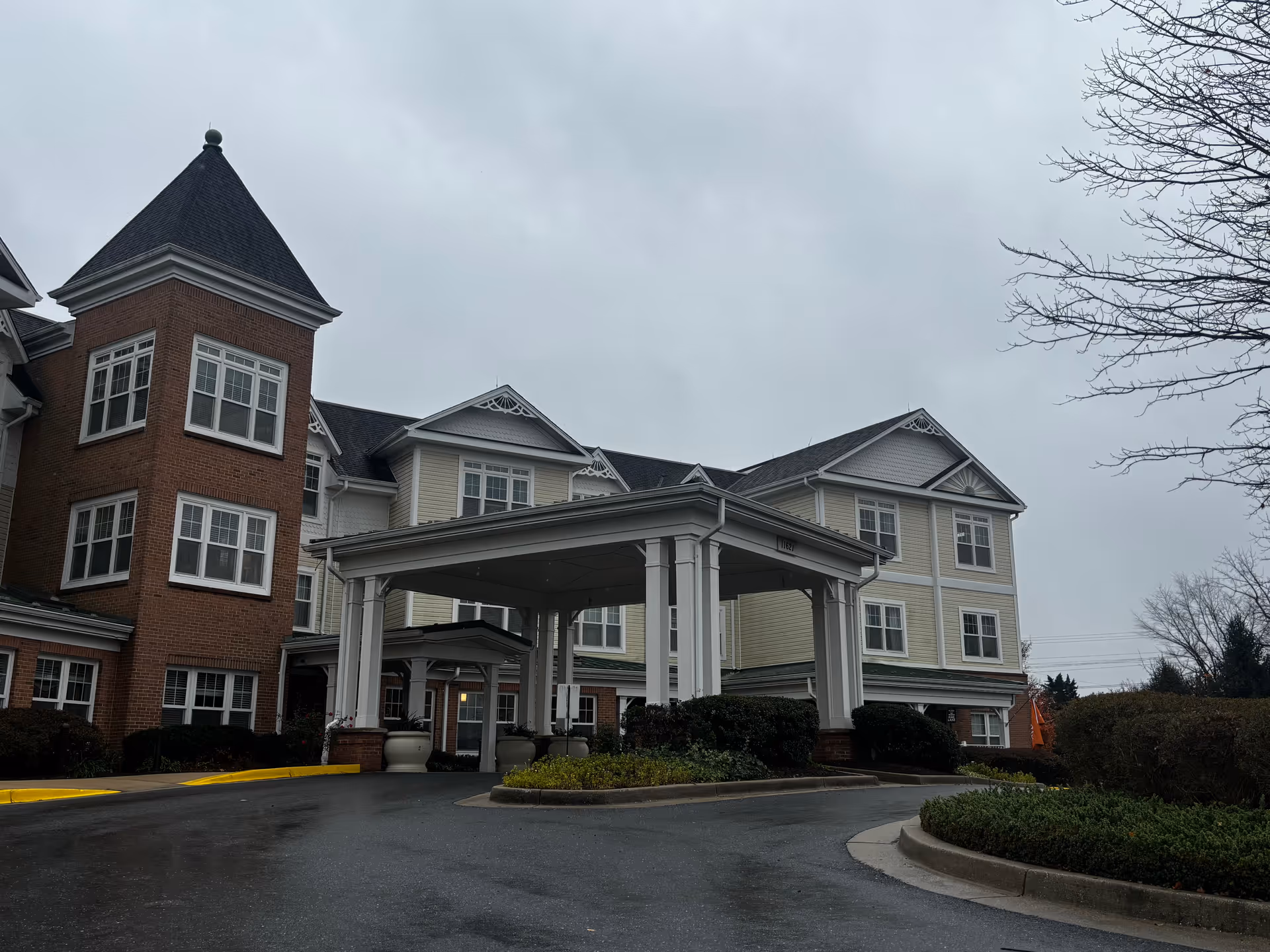 Front entrance of a multi-story senior living building with a covered porte-cochere and surrounding landscaping on an overcast day.