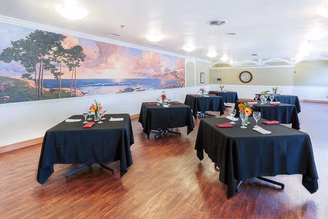 Dining room with several square tables covered with black tablecloths, each set with glassware, napkins, and small flower arrangements. The room has wooden flooring, light-colored walls, a large mural of a coastal landscape, and a clock on the far wall.