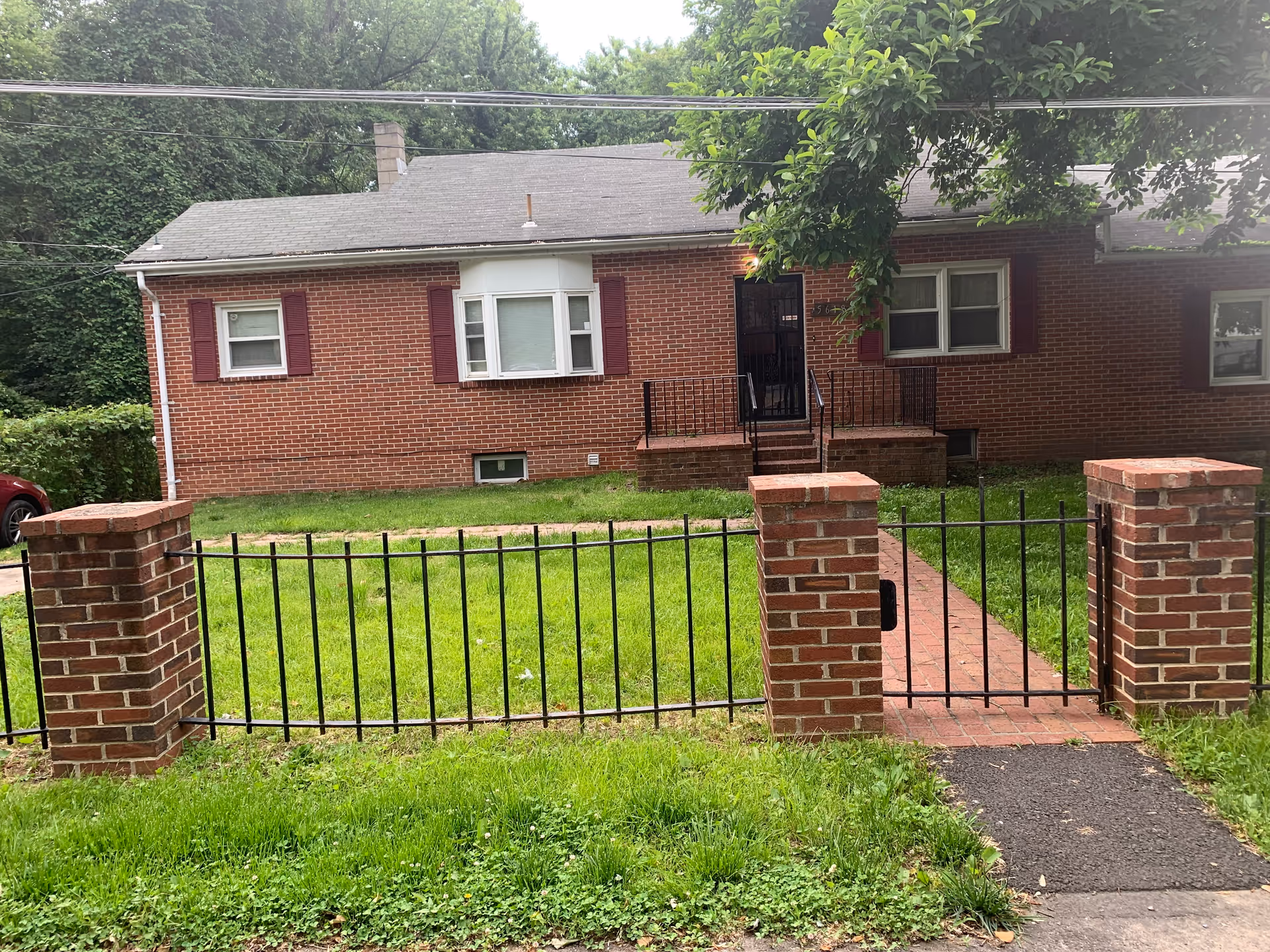 Front exterior view of a single-story brick house with a gray roof, three windows with red shutters, a black metal gate and fence supported by brick pillars, a small brick pathway leading to the front door, and green grass in the yard. Trees and bushes surround the property.