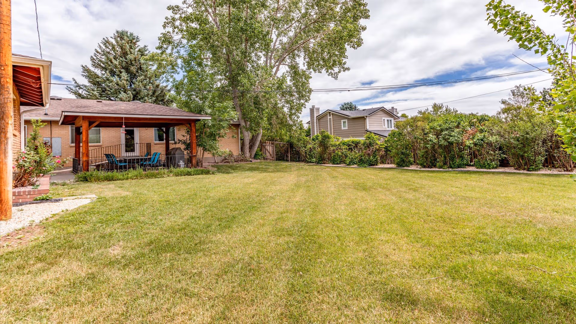 Large grassy backyard with a covered patio attached to a single-story building, trees, and neighboring houses in the background.
