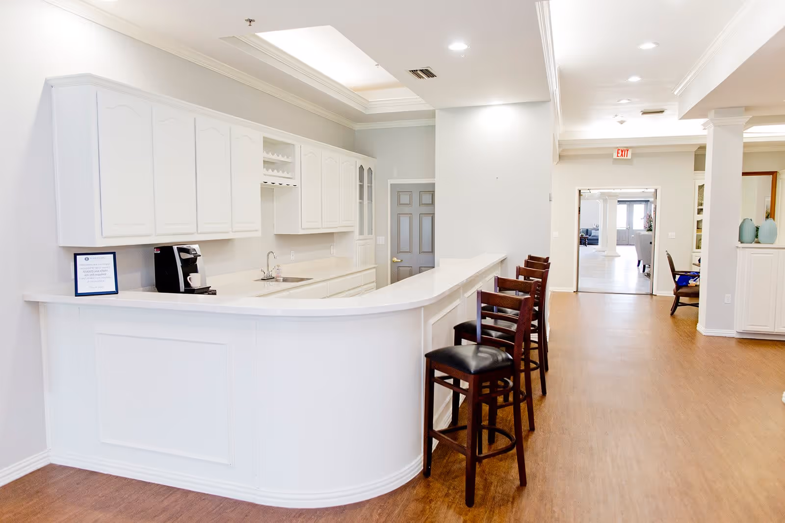 A bright and clean interior space featuring a long white counter with several dark wooden bar stools with black cushions. Behind the counter are white cabinets, a sink, and a coffee machine. The floor is wood, and the ceiling has recessed lighting. In the background, there is an open doorway leading to another room with more seating and natural light.