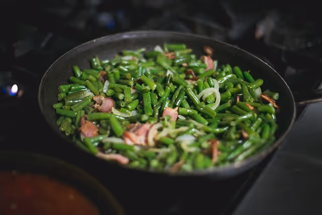 A frying pan on a stove containing cooked green beans mixed with pieces of bacon and sliced onions.