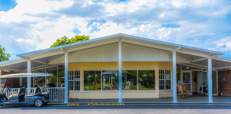 Front exterior view of a single-story building with a covered entrance supported by white columns. There are large windows with decorative star and hat decals, two rocking chairs near the entrance, and a parked golf cart on the left side. The sky is partly cloudy with blue patches.