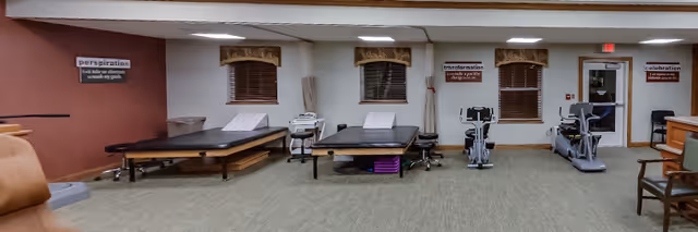 A therapy or rehabilitation room with two treatment tables, exercise equipment including a stationary bike and a treadmill, and motivational posters on the walls. The room has carpeted flooring, windows with brown valances, and a door leading outside or to another area.