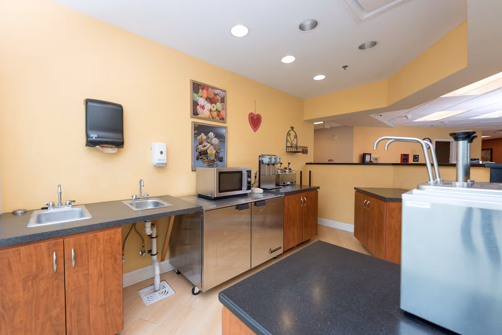 Small kitchenette with two sinks, countertop appliances including a microwave and beverage dispensers, stainless storage units and warm yellow walls.