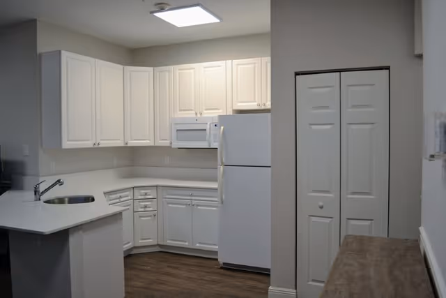 A clean, modern kitchen with white cabinets, a white refrigerator, a white microwave, and a white countertop with a built-in sink. The floor is wood, and there is a closet with double doors on the right side.