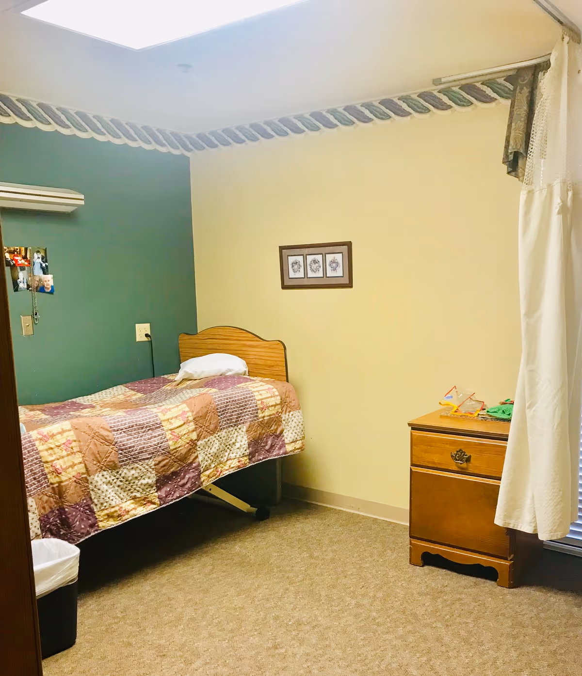 A small bedroom in a care facility with a single bed covered by a patchwork quilt. The bed has a wooden headboard and a white pillow. There is a wooden nightstand with a drawer next to the bed, holding some small items. The walls are painted green and beige with a decorative border near the ceiling. A curtain partially covers a window with blinds, and a few photos are pinned to the green wall above the bed.