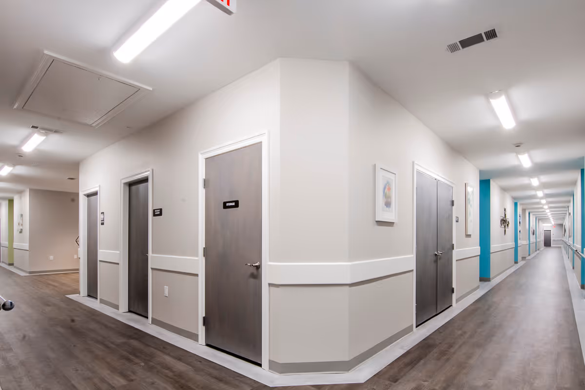 A clean, well-lit hallway in Shelton Manor Independent and Assisted Living with light-colored walls, wood flooring, and several closed doors labeled for storage and other rooms. The hallway has handrails along the walls and framed artwork hanging on the walls.