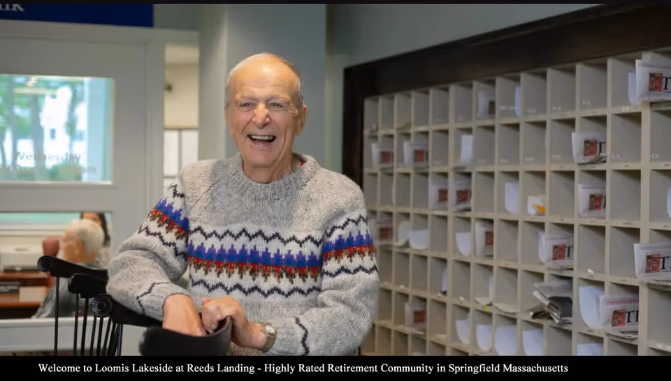An elderly man wearing a gray sweater with colorful patterns is smiling and leaning on the back of a black chair inside a room with a wall of mail slots filled with papers behind him.