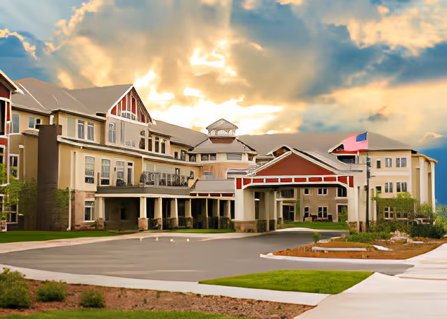 Exterior view of a large senior living facility building with multiple windows, a covered entrance, and an American flag flying on a flagpole. The sky is partly cloudy with sunlight breaking through.