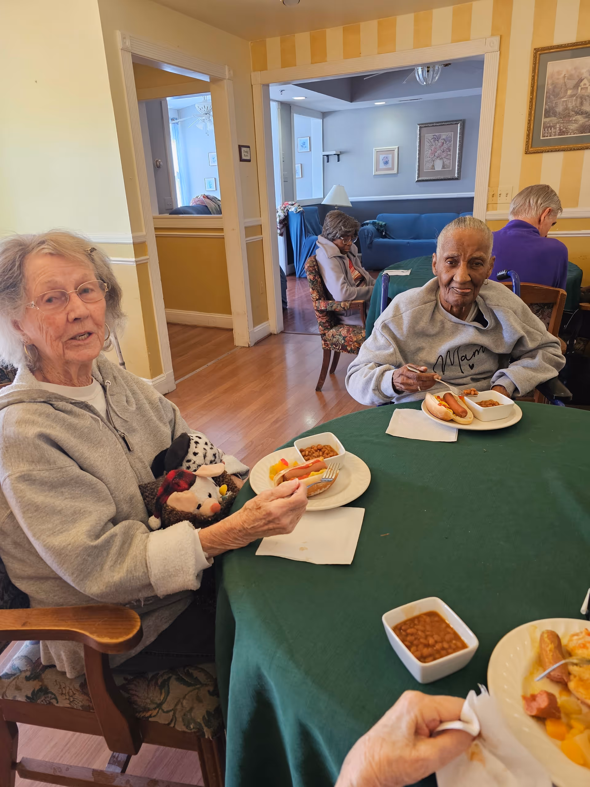 Several elderly individuals seated around a green tablecloth-covered table in a dining area, eating meals that include hot dogs, baked beans, and fruit. The room has yellow and white striped walls and wooden flooring, with a living room visible in the background.