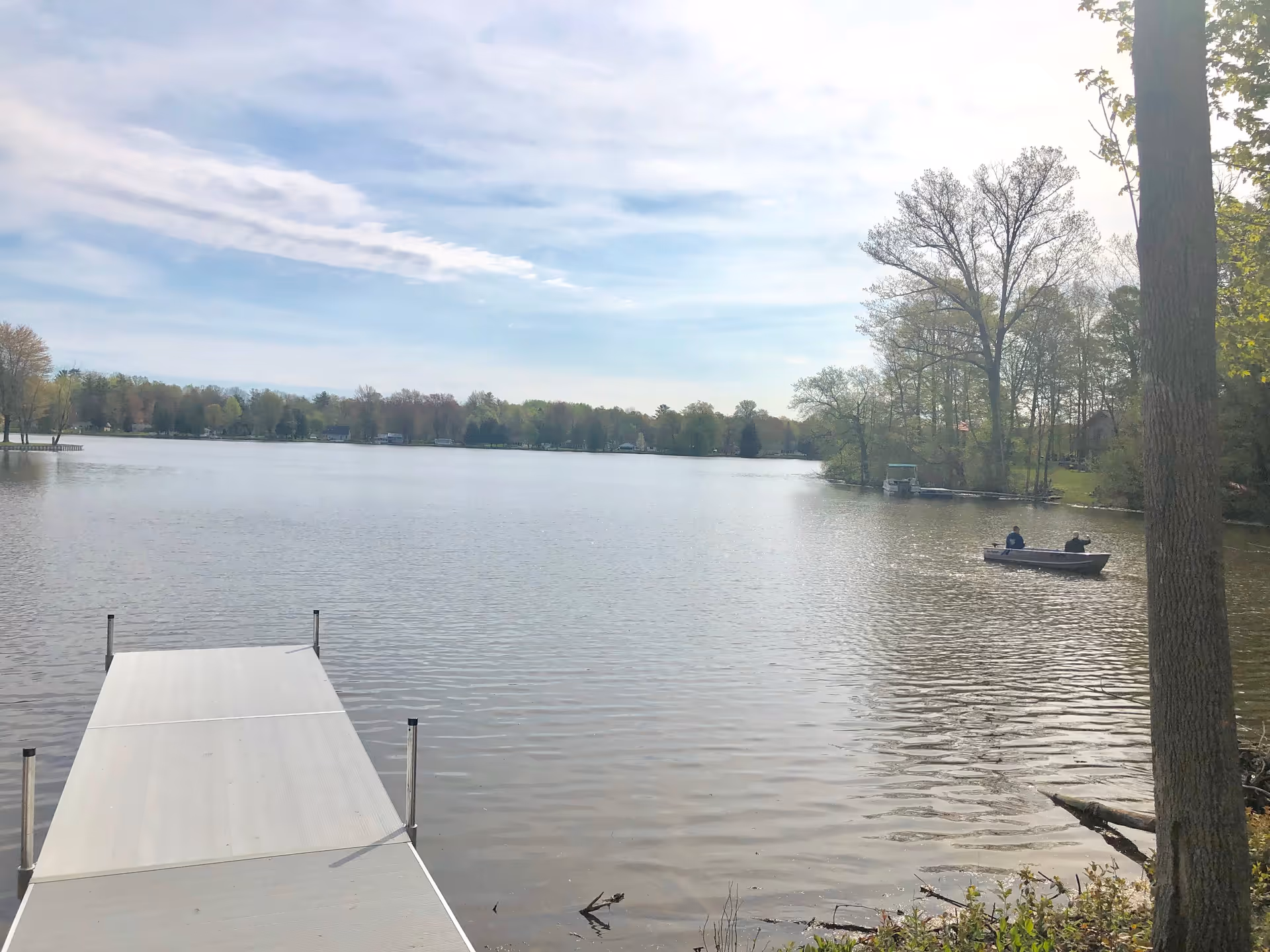 A peaceful lake scene with a dock extending into the water in the foreground. Two people are sitting in a small boat on the right side of the lake. Trees with early spring foliage surround the lake under a partly cloudy sky.