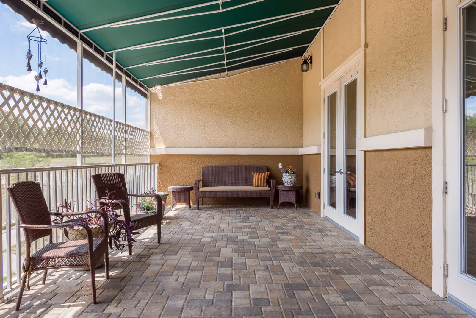 A covered outdoor patio area with a green awning overhead, two brown wicker chairs with plants beside them, a brown wicker loveseat with a striped pillow, two small round tables, and a potted flower arrangement. The patio has a brick floor and beige stucco walls with white trim. French doors lead inside the building.