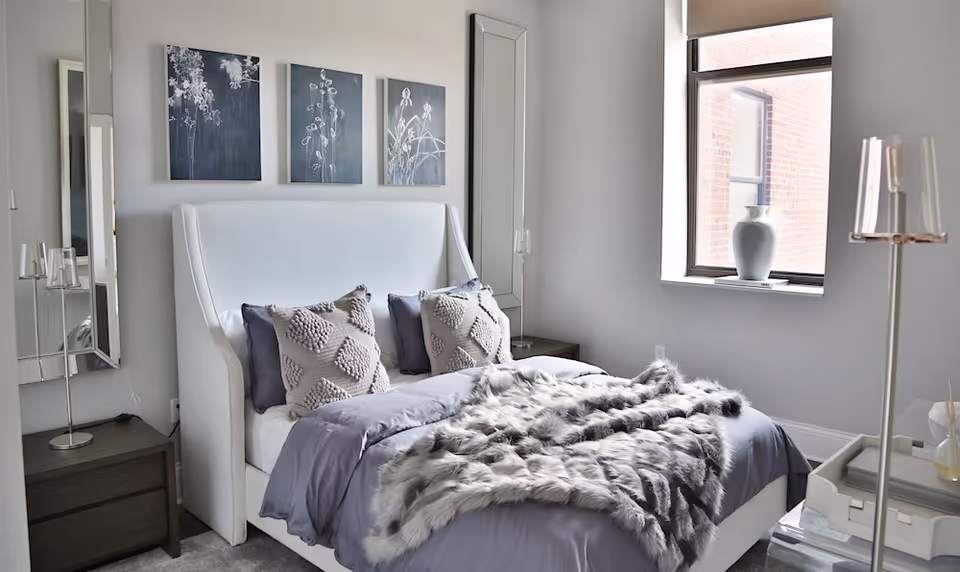 Light-filled modern bedroom with a white upholstered bed, gray bedding and a faux-fur throw, pillows, bedside tables, mirrors, artwork and a window.