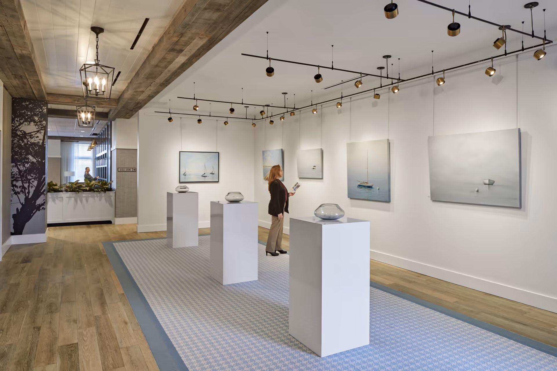An interior gallery space with wooden floors and white walls displaying paintings of boats and water scenes. Four white pedestals hold glass art pieces. A woman is standing and looking at the paintings while holding a booklet. The ceiling has track lighting and decorative hanging lights. In the background, a person is visible near a reception or seating area with plants.