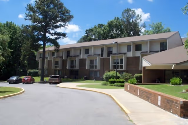 Exterior view of a two-story brick apartment building with several windows, surrounded by trees and greenery under a blue sky with some clouds. There are a few parked cars along the driveway leading to the building entrance.