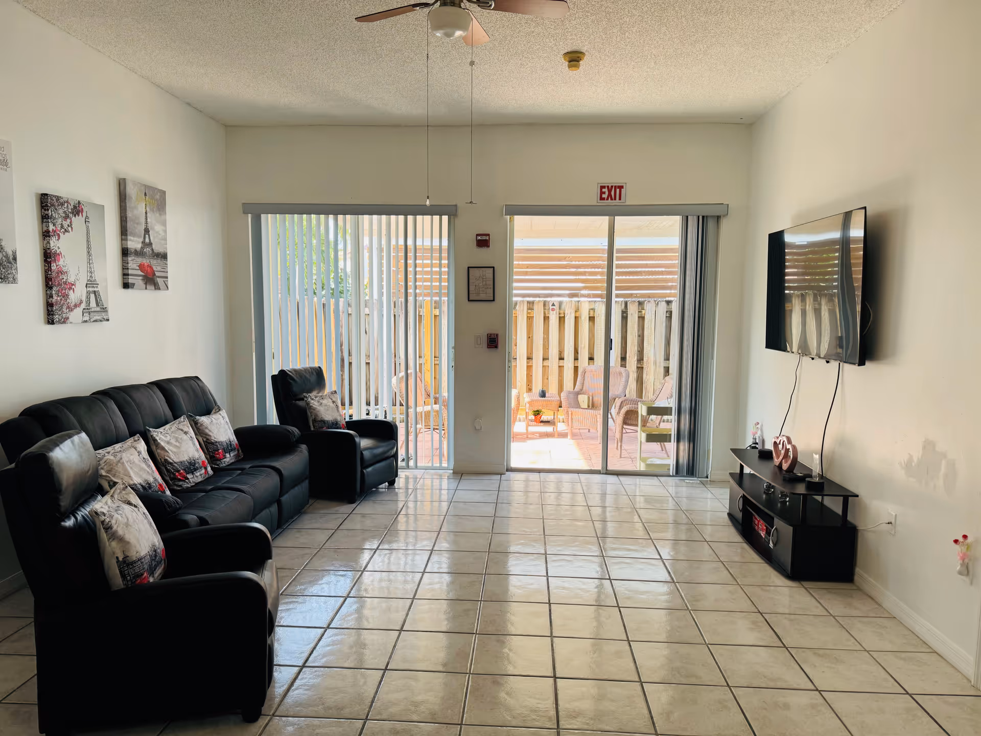 A living room with a black leather sofa and two matching armchairs, each with decorative pillows featuring Eiffel Tower designs. The room has tiled flooring, white walls with three framed pictures of the Eiffel Tower, a ceiling fan, and a wall-mounted flat-screen TV above a black TV stand. Two sliding glass doors with vertical blinds lead to an outdoor patio area with wicker chairs and a wooden fence. An exit sign is visible above one of the doors.