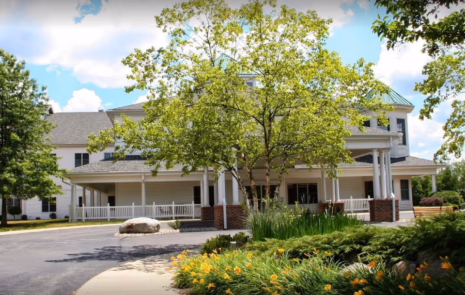 Front entrance of a large white senior living building with columns, a covered driveway, trees and flower beds in the foreground.