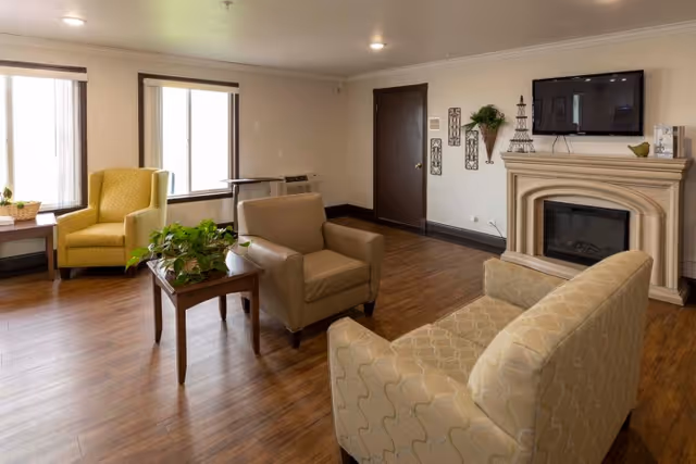 A bright common living room with sofas and armchairs around a small table, a wall-mounted TV and a fireplace.