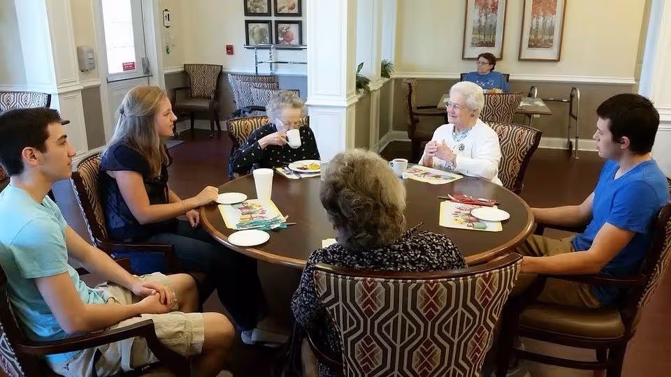 Several elderly residents and younger visitors sit around a round dining table in a senior living facility, chatting and having a meal.
