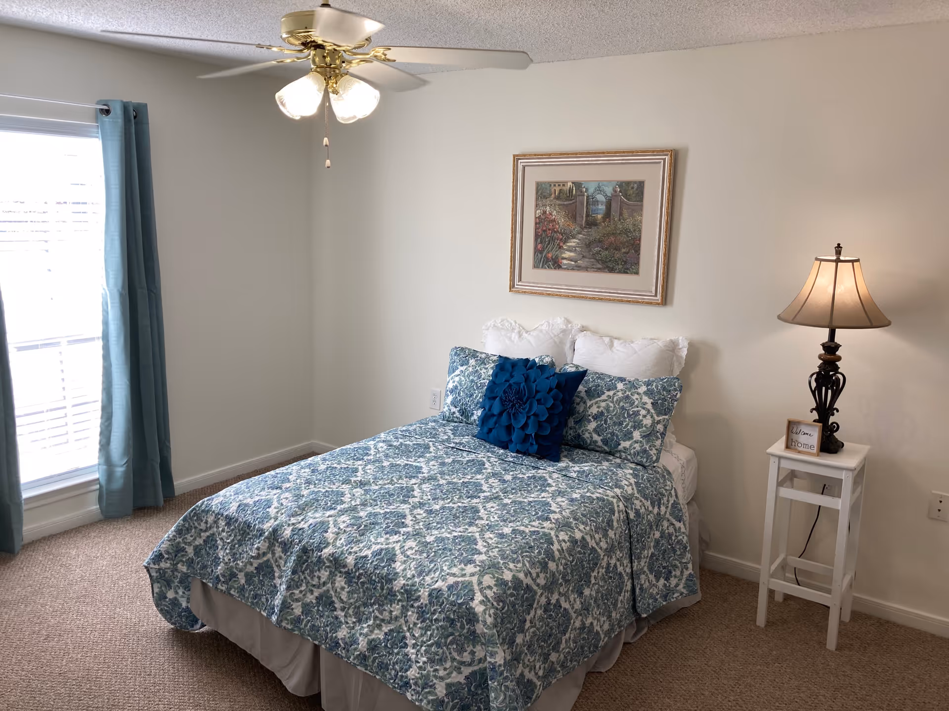 A bedroom with a bed covered in a blue and white patterned quilt and pillows, including a decorative blue flower-shaped pillow. There is a white nightstand with a lamp and a small sign that says 'Welcome Home'. A framed picture hangs on the wall above the bed. Blue curtains frame a window letting in natural light, and a ceiling fan with lights is mounted on the ceiling.