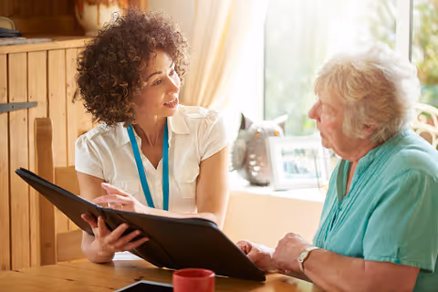 A caregiver or staff member with curly hair and a white shirt is sitting at a wooden table, holding a black folder and talking to an elderly woman with short gray hair wearing a teal top. They are in a well-lit room with a window and light-colored curtains in the background.