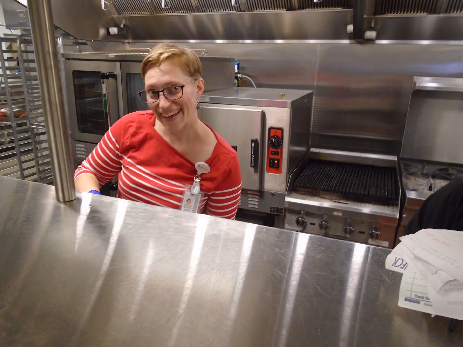 A smiling woman wearing glasses and a red and white striped shirt stands behind a stainless steel counter in a commercial kitchen with ovens and grills in the background.