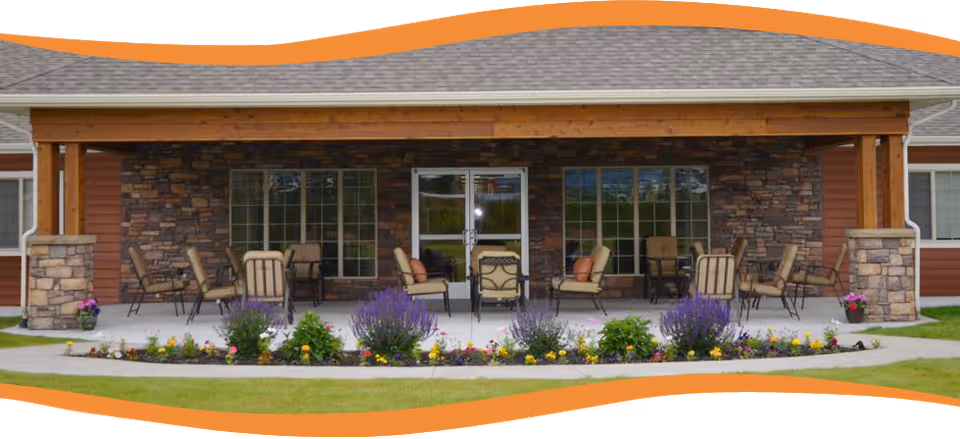 Outdoor covered patio area with multiple cushioned chairs and tables arranged on a concrete floor, surrounded by a flower bed with colorful flowers and green grass. The building behind has stone and wood siding with large windows and a glass door.
