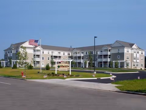 Exterior view of Cedar Creek Apartments II, a three-story residential building with beige siding and white trim. The building has multiple balconies and is surrounded by a well-maintained lawn and young trees. An American flag is flying on a flagpole near the entrance, and a sign with the facility name is visible in front of the building.