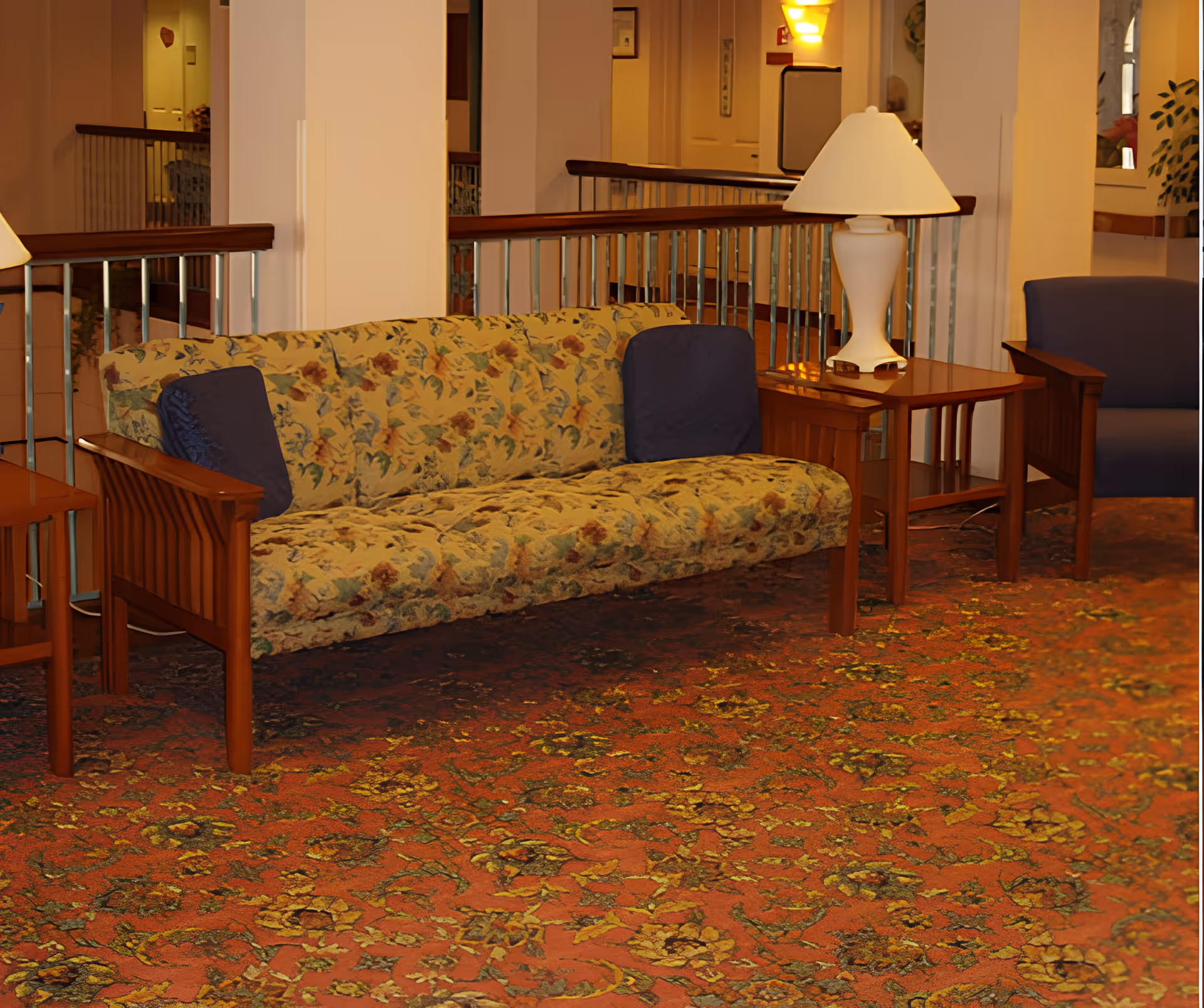 A floral-upholstered sofa with blue pillows, wooden side tables and lamps in a carpeted lounge area.
