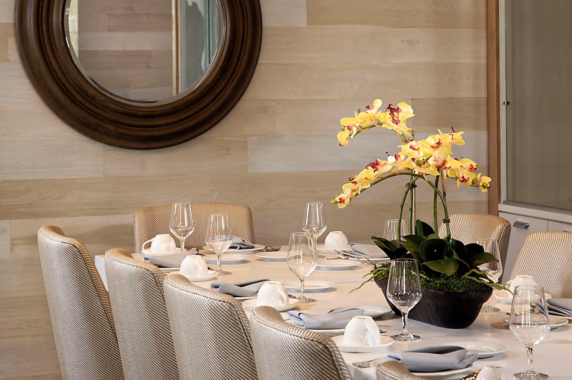 A dining table set for a meal with beige upholstered chairs around it, white plates, folded gray napkins, upside-down white cups, and empty wine glasses. A large black vase with yellow orchids is placed in the center of the table. A round wooden-framed mirror hangs on the light wood-paneled wall in the background.