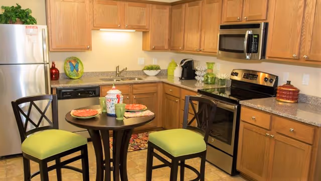 A modern kitchen with wooden cabinets, stainless steel refrigerator, oven, and microwave. There is a small round table with two chairs that have green cushions. The countertop has various decorative items including a bowl of greenery, a coffee maker, and colorful plates on the table.