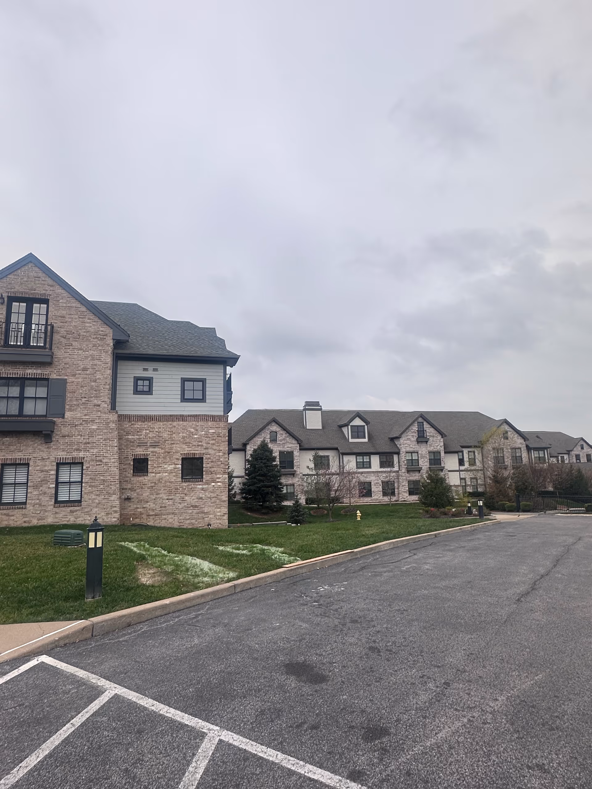 Exterior view of a senior living facility building with brick and siding facade, multiple windows, and a sloped roof under a cloudy sky. There is a paved parking area in the foreground and a grassy lawn with small trees and shrubs near the building.